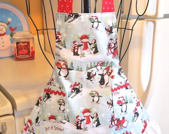 May include: A light blue apron featuring a repeating pattern of penguins playing in the snow, building snowmen, and skiing. The apron has red polka-dot neck straps, two front pockets with red trim, and a white eyelet lace ruffle at the hem.