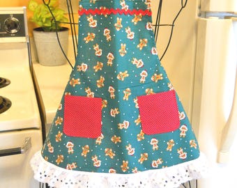May include: A teal apron featuring a repeating pattern of gingerbread people and white dots. The apron includes two red pockets with white polka dots, a red rickrack trim along the chest, and a white eyelet lace ruffle along the bottom hem.