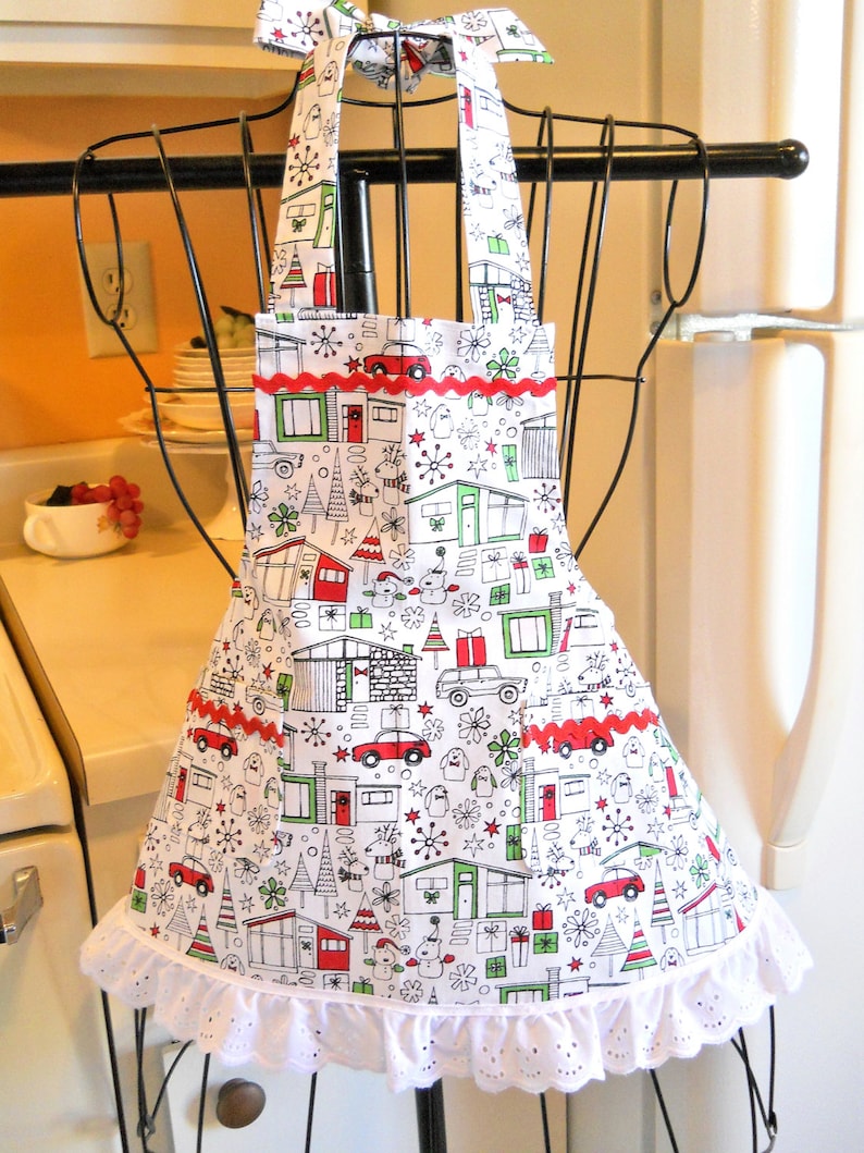 May include: A white apron featuring a repeating holiday pattern of mid-century modern houses, red cars, Christmas trees, and gifts. The apron includes two front pockets accented with red rickrack trim and a white eyelet lace ruffle at the hem.