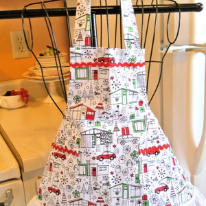 May include: A white apron featuring a repeating holiday pattern of houses, cars, gifts, and trees in red, green, and black. The apron includes a red rickrack trim across the chest and pockets, a white eyelet lace ruffle at the hem, and a bow.