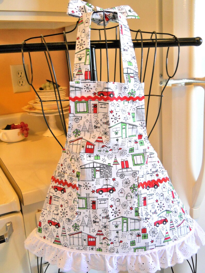 May include: A white apron featuring a repeating holiday pattern of houses, cars, gifts, and trees in red, green, and black. The apron includes a red rickrack trim across the chest and pockets, a white eyelet lace ruffle at the hem, and a bow.