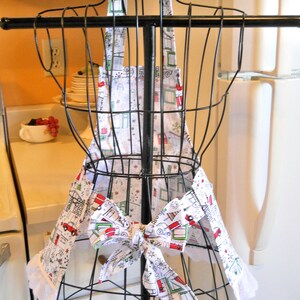 May include: A white apron featuring a repeating pattern of red and green holiday-themed campers, trees, and snowflakes. The apron includes a neck strap with a bow, a waist tie with a bow, and white eyelet lace trim along the bottom edges.