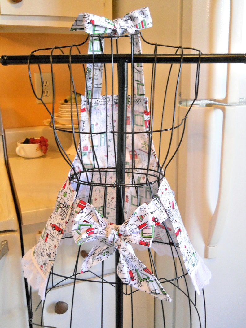 May include: A white apron featuring a repeating pattern of red and green holiday-themed campers, trees, and snowflakes. The apron includes a neck strap with a bow, a waist tie with a bow, and white eyelet lace trim along the bottom edges.