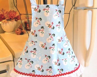 May include: A light blue apron featuring a repeating pattern of snowmen wearing black top hats and red scarves. The apron includes two front pockets, a red rickrack trim, and a white eyelet lace ruffle along the bottom hem with neck ties.
