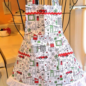 May include: A white apron featuring a repeating holiday print of mid-century modern houses, cars, gifts, and reindeer in red, green, and black. The apron includes two front pockets with red rickrack trim and a white eyelet lace ruffle at the hem.