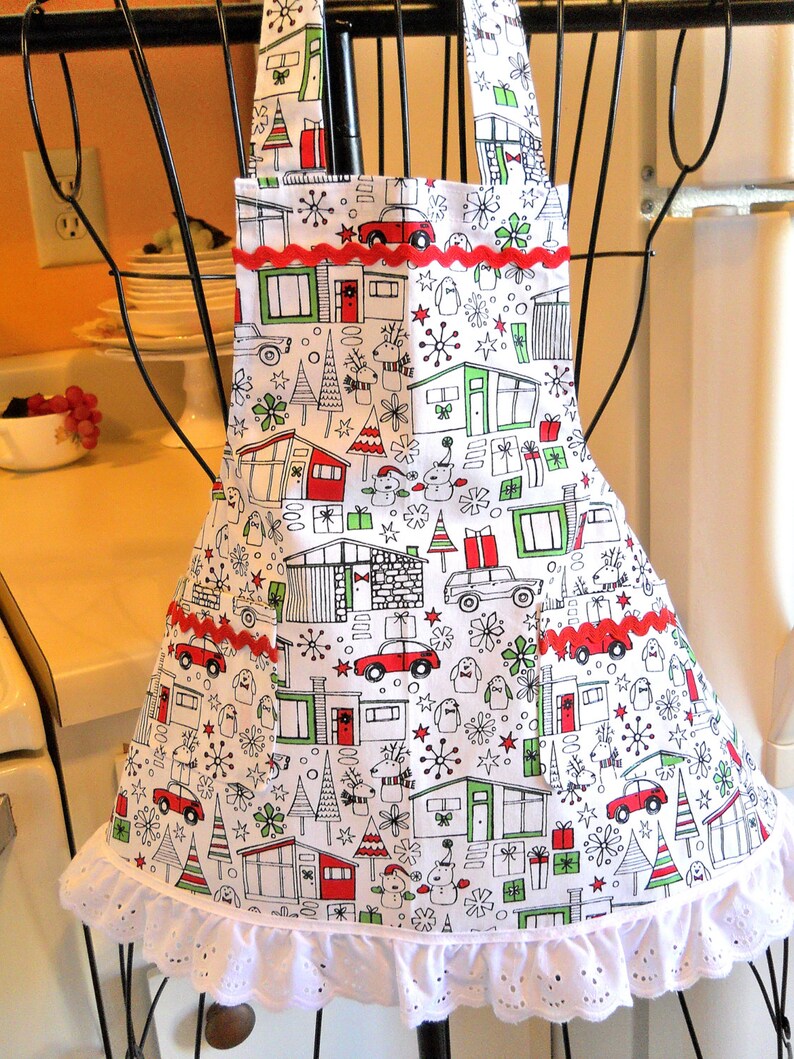 May include: A white apron featuring a repeating holiday print of mid-century modern houses, cars, gifts, and reindeer in red, green, and black. The apron includes two front pockets with red rickrack trim and a white eyelet lace ruffle at the hem.