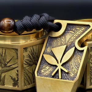 Peut inclure: Gros plan sur un pendentif doré avec un cordon noir. Le pendentif présente un motif de feuille stylisé et des motifs gravés complexes. Une perle en bois est attachée au cordon. Le pendentif est à côté d'une boîte dorée avec des motifs floraux gravés.