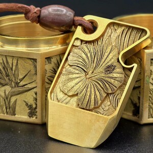 Peut inclure: Pendentif doré avec un motif de fleur d'hibiscus détaillé. Le pendentif rectangulaire aux bords arrondis est attaché à un cordon en cuir marron. En arrière-plan, une boîte dorée avec des motifs floraux gravés.