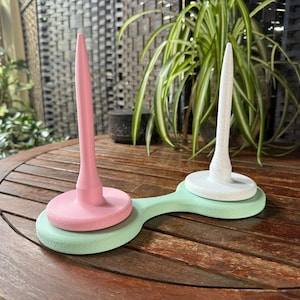 Op de afbeelding: Een tweedelige garenklos in pastelkleuren. De ene kant is roze, de andere wit, beide op een lichtgroene basis. De klos is ontworpen om garen vast te houden voor brei- of haakprojecten. De klos staat op een houten tafel.