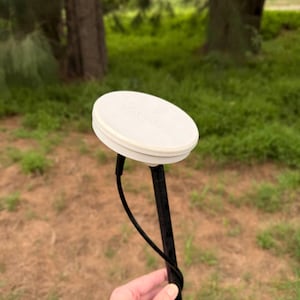 May include: A white, circular device with a black pole and a black cable. The device is held in a hand, with a blurred background of trees and grass. The device appears to be a surveying tool.