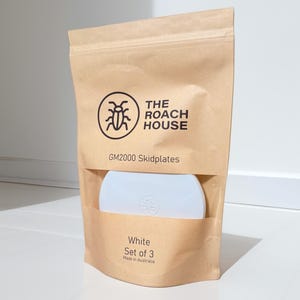 May include: A brown paper bag with a clear window displaying white skidplates. The bag has the text "THE ROACH HOUSE" and "GM2000 Skidplates". Below the window, the text reads "White Set of 3 Made in Australia".