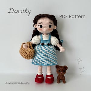 May include: A crocheted doll of Dorothy from The Wizard of Oz, wearing a blue and white dress, red shoes, and holding a basket. The doll has dark brown hair in pigtails. A small brown crocheted teddy bear is next to the doll. Text reads "Dorothy" and "PDF Pattern".
