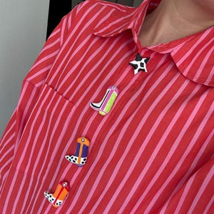 May include: A red and pink striped button-down shirt with decorative boot-shaped buttons in various colors. The buttons are in the shape of cowboy boots, with details like polka dots and stripes. A black and white star-shaped button is at the top.