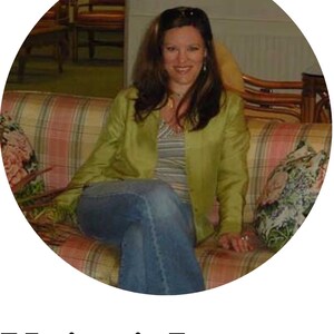 May include: A woman with long brown hair is sitting on a patterned couch. She is wearing a green jacket, a striped shirt, and blue jeans.
