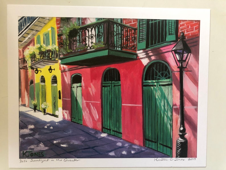 May include: A painting of a narrow street in New Orleans with colorful buildings and green doors. The buildings have balconies and are painted in shades of yellow, pink, and red. The street is paved with gray stones and there is a lamppost on the right side of the image. The painting is signed "K. Jones 2019" and titled "Sunlight in the Quarter".