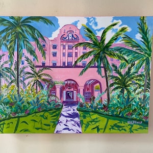May include: A painting depicting a pink building with arched doorways and palm trees in front. The building has a tiled roof and white trim. The palm trees are green and have long fronds. The painting is signed "K. Jones" in the bottom right corner.