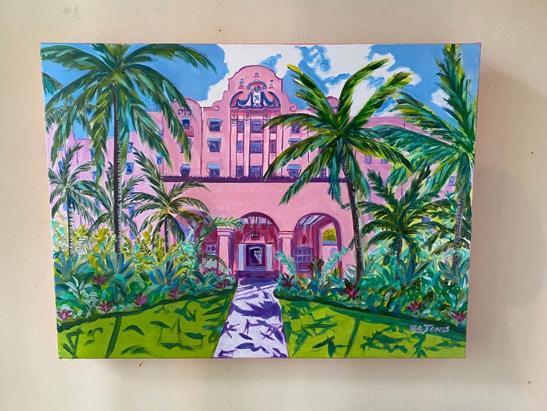 May include: A painting depicting a pink building with arched doorways and palm trees in front. The building has a tiled roof and white trim. The palm trees are green and have long fronds. The painting is signed "K. Jones" in the bottom right corner.