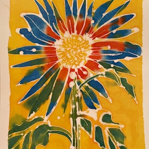May include: A colorful batik artwork of a sunflower. The flower has blue, red, and white petals with a yellow center. The stem and leaves are green. The background is yellow. The text "Sunflower Blue" is visible.