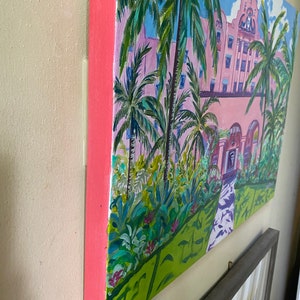 May include: A painting depicting a pink building with palm trees in front. The building has white trim and a clock tower. The palm trees are green with brown trunks. The painting is on a canvas with a pink frame.