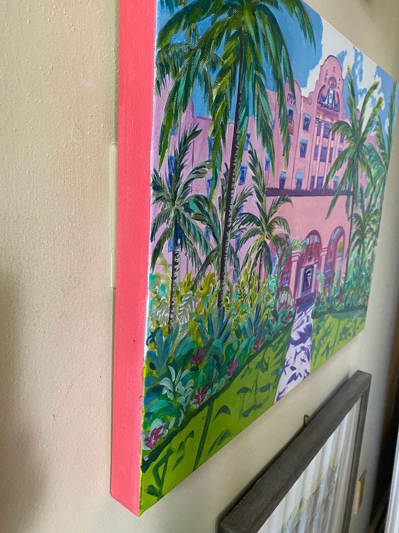 May include: A painting depicting a pink building with palm trees in front. The building has white trim and a clock tower. The palm trees are green with brown trunks. The painting is on a canvas with a pink frame.