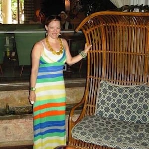May include: A woman wearing a colourful striped maxi dress stands next to a large, brown wicker chair with a patterned cushion. The chair has a high back and a curved shape.