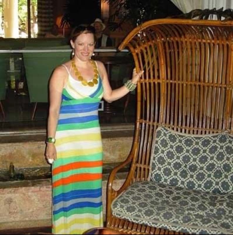 May include: A woman wearing a colourful striped maxi dress stands next to a large, brown wicker chair with a patterned cushion. The chair has a high back and a curved shape.