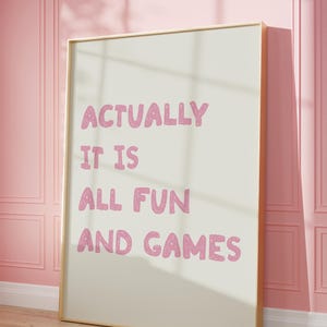 May include: A framed print with the words "ACTUALLY IT IS ALL FUN AND GAMES" in pink lettering. The print has a cream-colored background and a gold frame. It leans against a pink wall with a wooden floor.