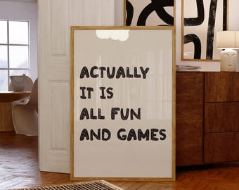 May include: A framed print with the text "ACTUALLY IT IS ALL FUN AND GAMES" in black letters. The print is in a room with wooden floors and a wooden cabinet. Two other framed abstract art pieces hang on the wall.