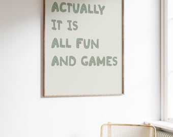 May include: A framed wall art print with the text "ACTUALLY IT IS ALL FUN AND GAMES" in green letters. The print is in a light-colored frame and hangs on a white wall. A gold-framed chair with a woven back and seat is below the print.