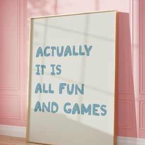 Könnte beinhalten: Gerahmtes Poster mit dem Text "ACTUALLY IT IS ALL FUN AND GAMES" in blauer Schrift. Das Poster lehnt an einer rosa Wand mit Holzboden. Der Rahmen hat eine hellbraune Farbe.