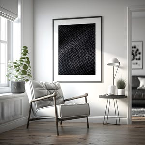 May include: A contemporary living room scene. A framed black and white artwork with a textured design hangs on the wall. A white armchair with wooden arms is in the foreground. A side table with a lamp and plant is also present.
