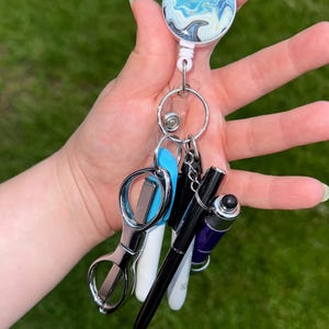 Puede incluir: Un portatarjetas con un diseño veteado azul y blanco, sujeto a un llavero con varios accesorios. Los accesorios incluyen unas tijeras plateadas, bolígrafos y un pequeño bolígrafo morado.
