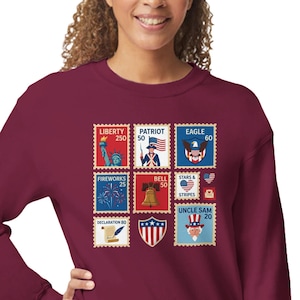 May include: A maroon sweatshirt with a collage of colourful postage stamp designs. The stamps show patriotic symbols like the Statue of Liberty, an eagle, fireworks, the Liberty Bell, and Uncle Sam. The sweatshirt is a comfortable, casual style.