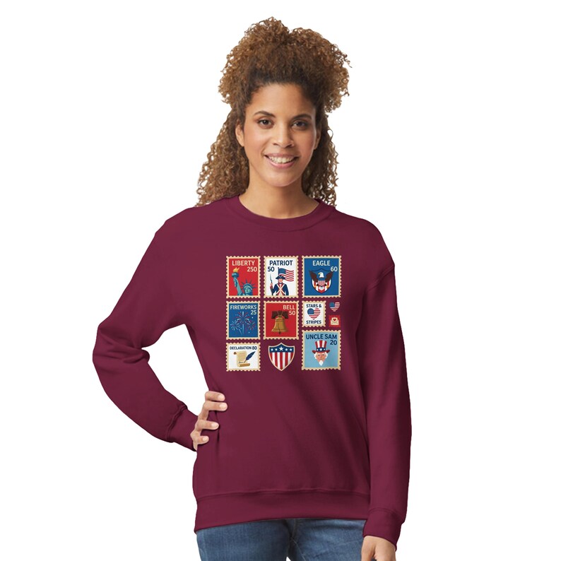 May include: A maroon sweatshirt with a collage of colourful postage stamp designs. The stamps show patriotic symbols like the Statue of Liberty, an eagle, fireworks, the Liberty Bell, and Uncle Sam. The sweatshirt is a comfortable, casual style.