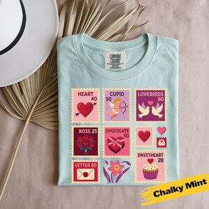 Op de afbeelding: Een lichtblauw t-shirt met Valentijnsdag-thema postzegelontwerpen. De postzegels tonen harten, Cupido, tortelduifjes, rozen, chocolade en een cupcake. Het shirt is gelabeld "Chalky Mint".
