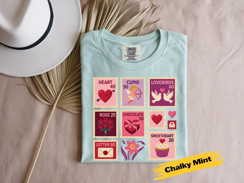 Op de afbeelding: Een lichtblauw t-shirt met Valentijnsdag-thema postzegelontwerpen. De postzegels tonen harten, Cupido, tortelduifjes, rozen, chocolade en een cupcake. Het shirt is gelabeld "Chalky Mint".