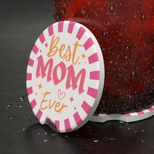 以下が含まれることがあります： ピンクと白のストライプのボーダーと、ピンクとオレンジ色の「Best MOM Ever」という文字が入った白い車のコースター。 コースターは、結露したグラスの隣にあります。