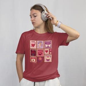 Op de afbeelding: Een bordeauxrood T-shirt met een reeks postzegelontwerpen met Valentijnsdagthema. De postzegels tonen harten, Cupido, tortelduifjes, rozen, chocolade en een cupcake. Elke postzegel heeft een nummer. Het T-shirt wordt gedragen.