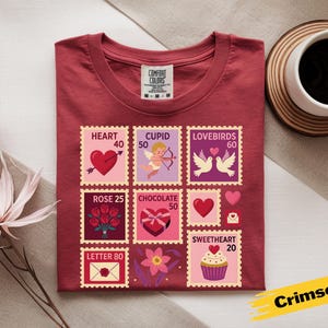 Op de afbeelding: Karmizijnrood T-shirt met Valentijnsdag postzegelontwerpen. De postzegels tonen harten, Cupido, tortelduifjes, rozen, chocolade en een cupcake. Het shirt bevat de tekst "Comfort Colors" en de kleurnaam "Crimson".