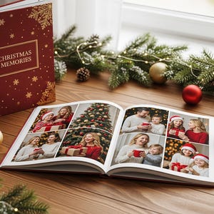 May include: A red Christmas Memories photo album with gold snowflake and floral accents, open to display family photos. A cup of coffee, ornaments, and greenery decorate the wooden table.