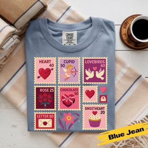 Op de afbeelding: Een blauw Comfort Colors t-shirt met Valentijnsdag postzegelontwerpen. De postzegels tonen harten, Cupido, tortelduifjes, rozen, chocolade en cupcakes. Een koffiekopje en opgevouwen dekens staan op de achtergrond.