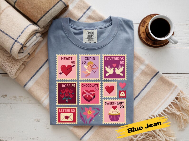 Op de afbeelding: Een blauw Comfort Colors t-shirt met Valentijnsdag postzegelontwerpen. De postzegels tonen harten, Cupido, tortelduifjes, rozen, chocolade en cupcakes. Een koffiekopje en opgevouwen dekens staan op de achtergrond.