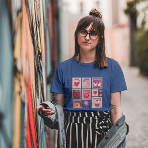 Op de afbeelding: Een blauw t-shirt met een reeks postzegelontwerpen met Valentijnsdagthema. De postzegels tonen harten, Cupido, tortelduifjes, rozen, chocolade en cupcakes. Het t-shirt wordt gedragen door een persoon met een bril.