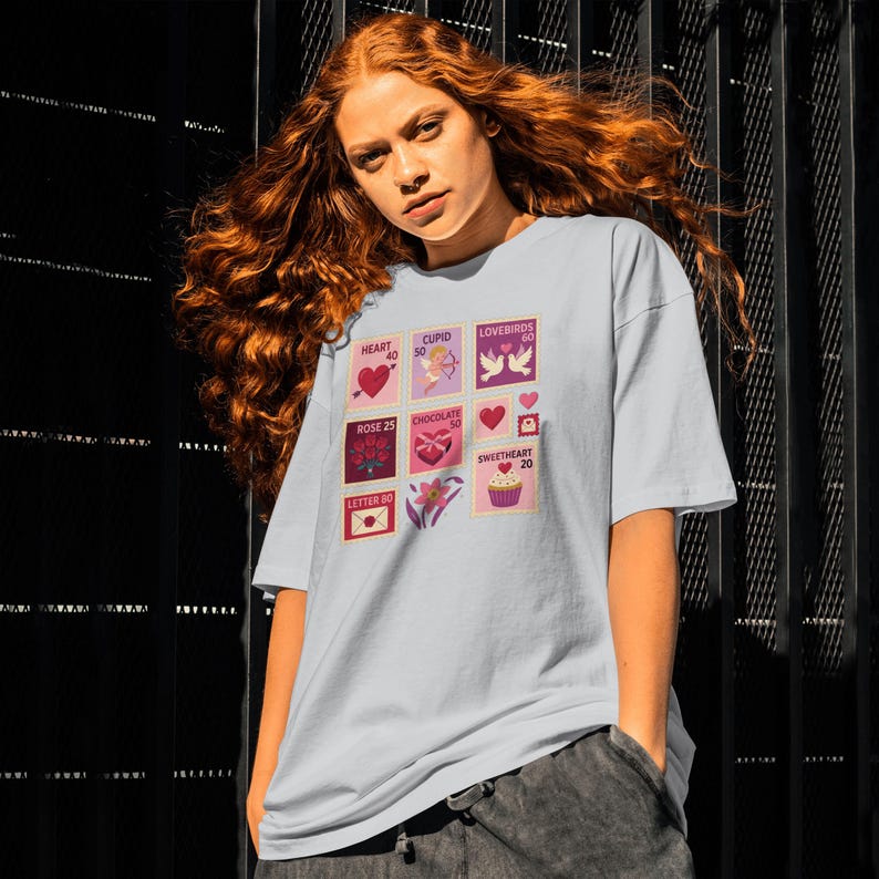 Op de afbeelding: Lichtblauw T-shirt met een collage van Valentijnsdag-postzegelontwerpen. De postzegels tonen harten, Cupido, tortelduifjes, rozen, chocolade en een cupcake. De persoon die het T-shirt draagt heeft rood haar.