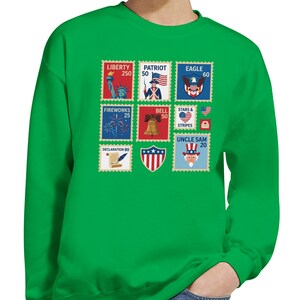 May include: Green sweatshirt with a series of patriotic stamp designs. The stamps show the Statue of Liberty, an American flag, an eagle, fireworks, a bell, stars and stripes, and Uncle Sam. The sweatshirt is made of a soft material.