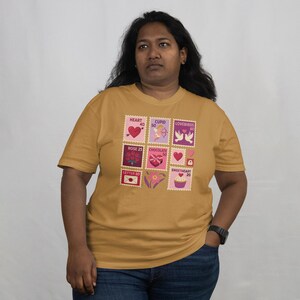 Op de afbeelding: Mosterdgeel T-shirt met Valentijnsdag postzegelontwerpen. De postzegels tonen harten, Cupido, tortelduifjes, rozen, chocolade en een cupcake. Het shirt is een T-shirt met korte mouwen en een ronde hals.