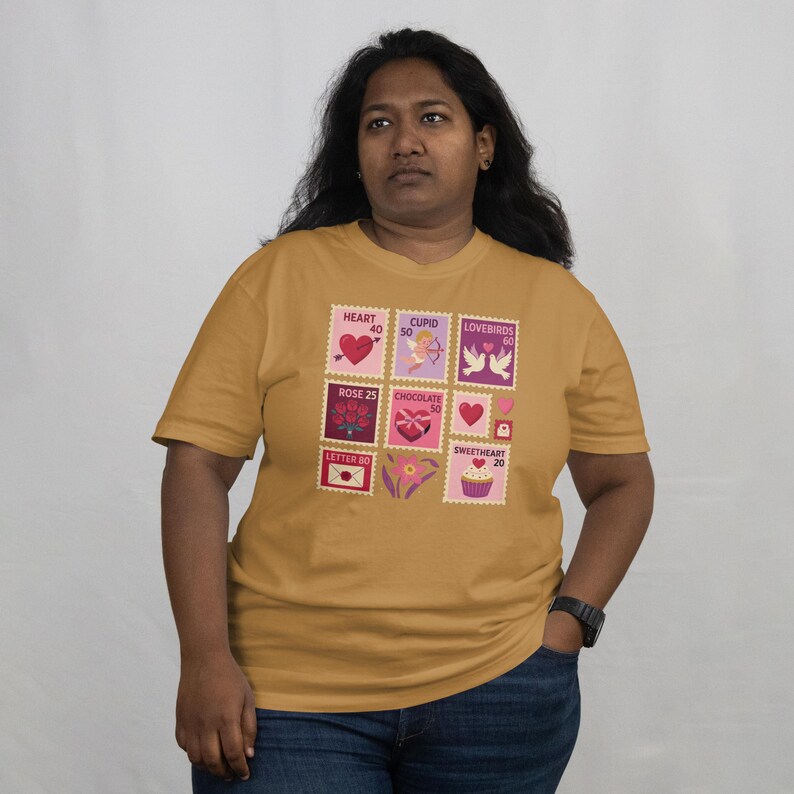 Op de afbeelding: Mosterdgeel T-shirt met Valentijnsdag postzegelontwerpen. De postzegels tonen harten, Cupido, tortelduifjes, rozen, chocolade en een cupcake. Het shirt is een T-shirt met korte mouwen en een ronde hals.