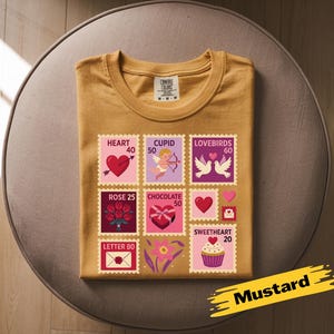Op de afbeelding: Mosterdkleurig T-shirt met een Valentijnsdag stempelontwerp. De postzegels bevatten harten, Cupido, tortelduifjes, rozen, chocolade en een cupcake. Het shirt is op een ronde, bruine ondergrond gevouwen.