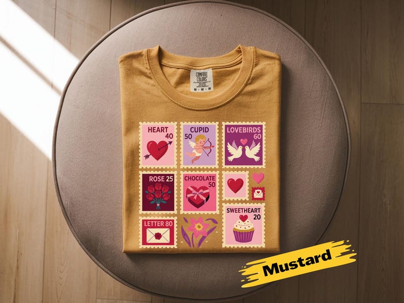 Op de afbeelding: Mosterdkleurig T-shirt met een Valentijnsdag stempelontwerp. De postzegels bevatten harten, Cupido, tortelduifjes, rozen, chocolade en een cupcake. Het shirt is op een ronde, bruine ondergrond gevouwen.