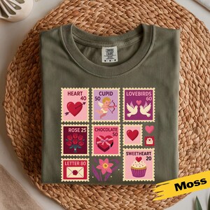 Op de afbeelding: Olijfgroen t-shirt met een Valentijnsdag stempelontwerp. De postzegels bevatten harten, Cupido, tortelduifjes, rozen, chocolade en een cupcake. Het woord "Moss" is in het geel geschreven.
